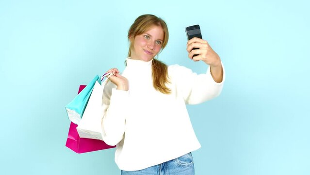 Young pretty blonde woman holding shopping bags while making a selfie isolated on background