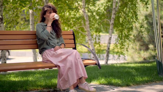 A girl in a pink skirt in the park on a bench drinks coffee.
