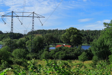 Power distribution lines. Electricity transfer. High voltage. Green summer nature with blue sky and some clouds. Colorful photo. House with brick roof hiding in the trees. Stockholm, Sweden, Europe.