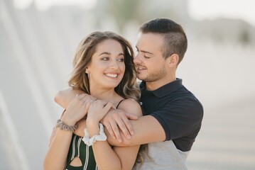 A close photo of a guy who is hugging his girlfriend in the modern urban space at the sunset in a Spain town. A couple of tourists on a date in the evening in Valencia.