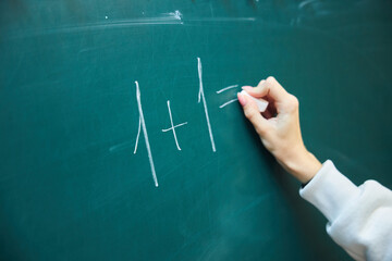 A teenager's hand with chalk writes a simple mathematical example on a green blackboard. The concept of education and training.