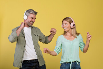 Enjoy music together. Happy middle aged spouses in wireless headphones listening songs and dancing, yellow background