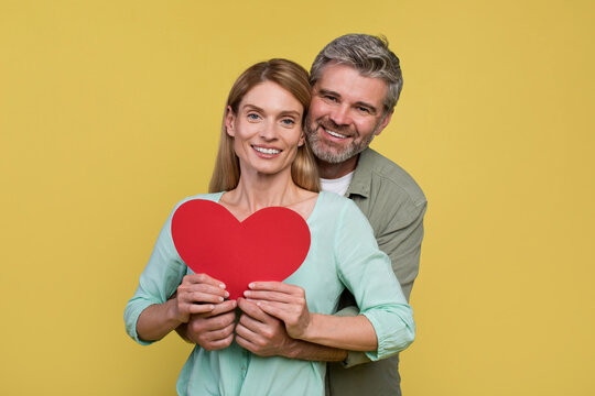 Loving middle aged spouses holding paper heart, man embracing wife brom back and smiling together at camera
