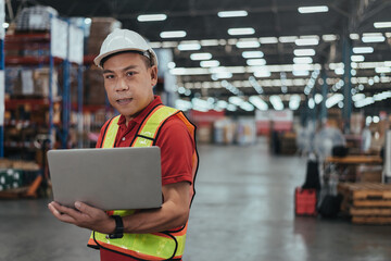 Warehouse Workers Standing and Holding Laptop in Logistic center. Asian Male Manager wearing safety vest and Hard Hat to working about shipment in storehouse, Working in Storage Distribution Center.