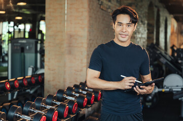 Portrait Asian man trainer using tablet in weights room at fitness gym. Workout training in fitness...