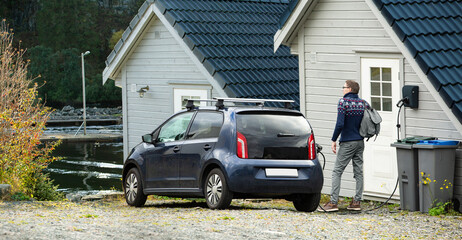 A man with a backpack next to charging electric car near a country house