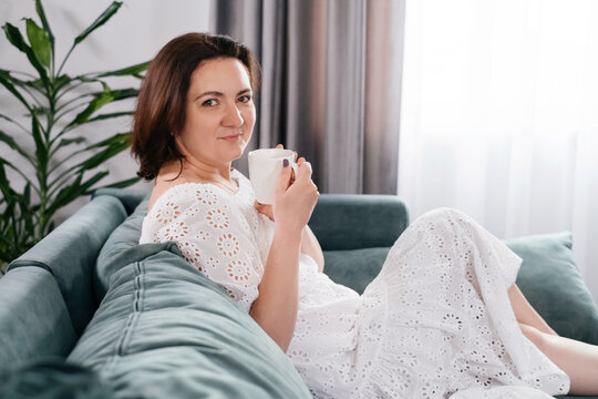 Woman Drinking Mug Of Coffee In Cozy Home Atmosphere In The Morning. Real Middle Age Plus Size Female In White Dress Relaxing On The Sofa, Modern House. Sunny Day With Cup Of Tea, Laying On Couch