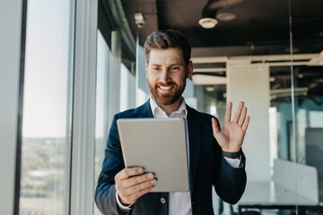 Happy caucasian businessman in suit working in office, having video chat with business partners via digital tablet