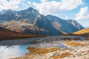 Beautiful mountain lake in hanging valley in orange autumn colors with view to large mountain range in sunlight. Lovely autumn landscape with small lake and high mountain top under clouds in blue sky.