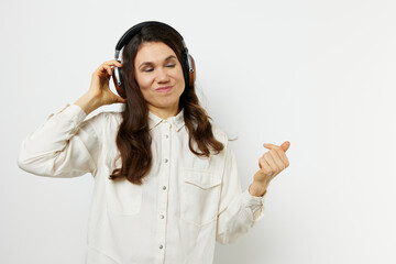 funny woman in light shirt stands listening to music in her headphones, closing her eyes with pleasure and holding headphones with her hands. Horizontal photo on a light background with an empty space