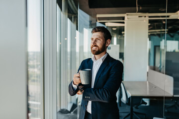 Handsome successful businessman having coffee break, standing next to window in his office with cup of coffee
