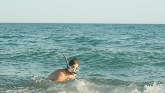 A man in a snorkeling mask looks suspiciously at the camera and sidles into the sea and dives. Slow mo