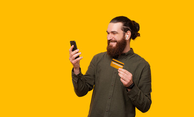 Naklejka premium Photo of a man making happily online shopping, holding a phone, credit card and sitting with his earbuds on