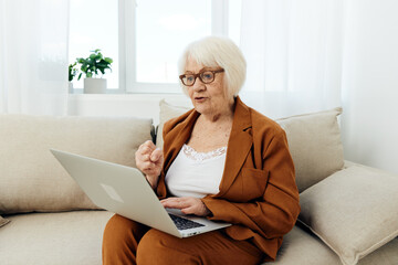 an evil, harmful old woman in a classic brown suit talks via video link holding a laptop on her lap sitting on the couch and holding her hand clenched in her fist, menacingly looking at the monitor