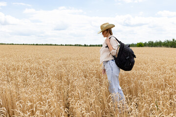 Naklejka na ściany i meble Back view of female agronomy with travel backpack walking at path in middle of wheat fields exploring international agriculture cultivation, tourist with backpack visiting gold plantation