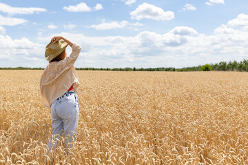 Naklejka na ściany i meble Back view of woman in big hat walking on wheat field in summer at ripe wheat ears. girl enjoying peacefulness and beautiful nature.