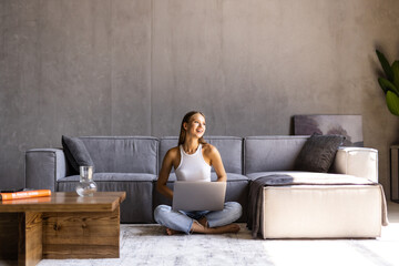 Portrait of a cheerful young woman listening to music with headphones and using laptop while leaning on a couch at home