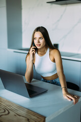 Young woman sits at the kitchen table using a laptop and talking on a phone