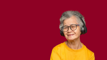 Cheerful elderly Asian woman with short gray hair wearing wireless headphones and looking at the digital camera with a smile while standing on a red background