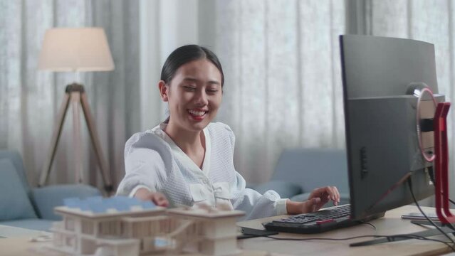 Asian Woman Engineer Holding And Looking At The Small House Model With Solar Panel Before Comparing It To The Pictures On A Desktop At Home
