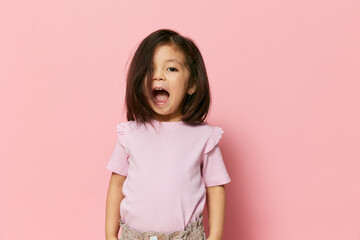 a cute little preschool girl on a pink background in a pink T-shirt is screaming loudly and waving...