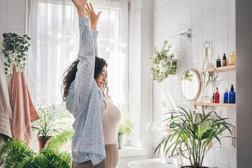 Young overweight woman stretching in light bathroom with minimalistic eco-flriendly design. White tile wall with shadows from the window. Wellness and body positivity concept