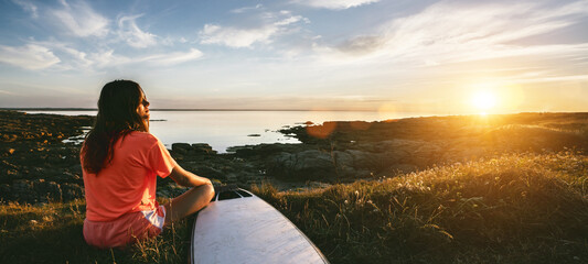 young surfer girl sitting at the ocean coast near her surfboard, watching the sunset and enjoy the...
