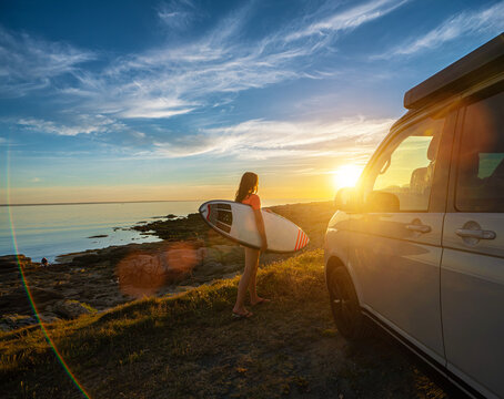 Surfer girl walking near her mini van and looking on the ocean at summer sunset  with a surfboard on her side