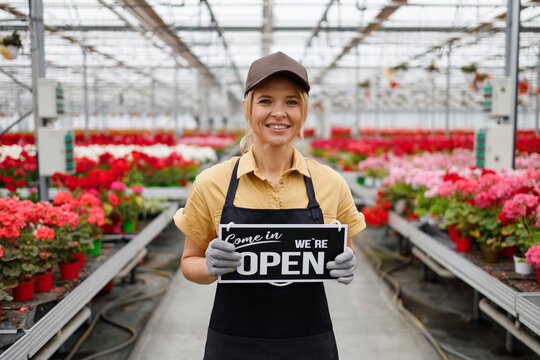 Portrait of pretty female flower greenhouse worker showing sign we are open