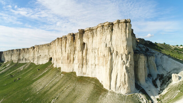 Bottom view of the high white rock on blue cloudy sky background. Shot. Sunny day and beautiful green meadow at the foot of the mountain, beauty of nature.