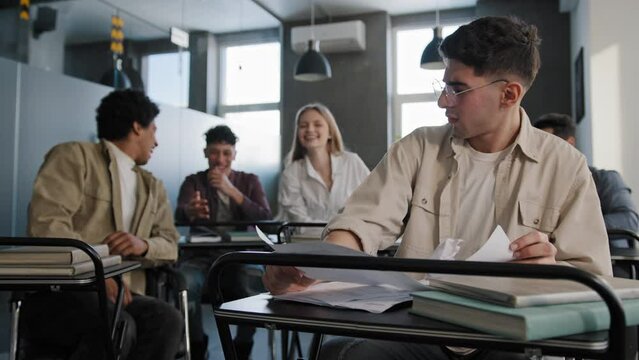 Sad worried upset disappointed caucasian student nerd wearing glasses sits in classroom at desk alone prepares for lesson reads notes aggressive angry classmates gossiping laughing bullying poor guy
