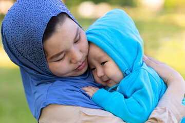 Cute young islamic kazakh woman hugging her little crying son, newborn boy. Yappy asian mother in hijab soothes, holding baby in hands outdoors. Family concept