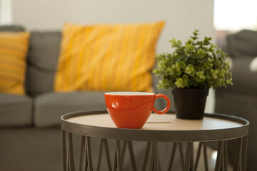 orange cup on table near couch. porcelain mug of aromatic coffee. background of gray sofa wit pillow