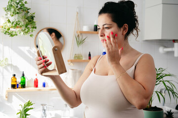 Young overweigh woman looking looking at the mirrow and examine her face. Wellness and body positivity