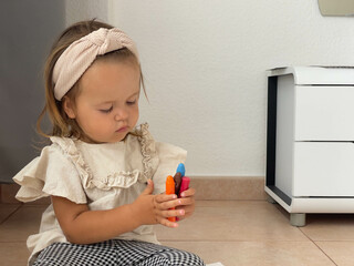 A baby girl is looking at the colourful chalk pastels while drawing a picture sitting on the floor near the window. 