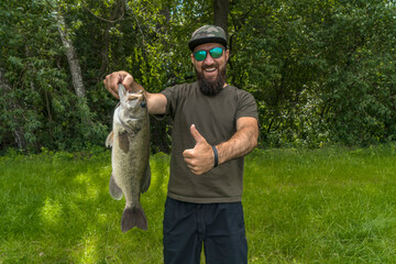 Successful bass fishing. Smiling bearded fisherman in sunglasses with bass fish show thumb up
