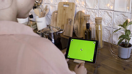 Rear view of a woman tapping on a tablet with green screen in the kitchen at home for recipes....