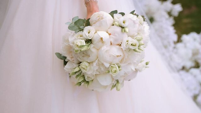 The bride in a lush dress holds a beautiful delicate bouquet of fresh flowers in her hand. The florist used peonies, eustomas and greenery in the bouquet