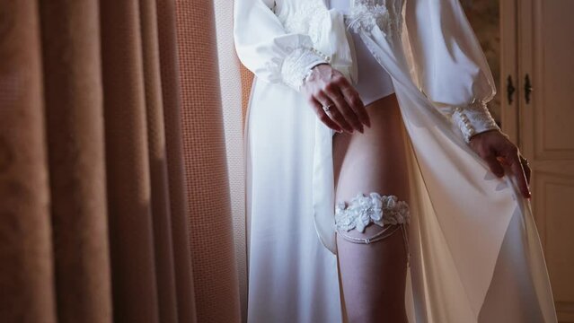 Caucasian sexy girl stands and opens her silk robe and exposes her leg. Slim woman with a garter on her leg. Wedding detail. Morning bride. Preparation for the wedding ceremony