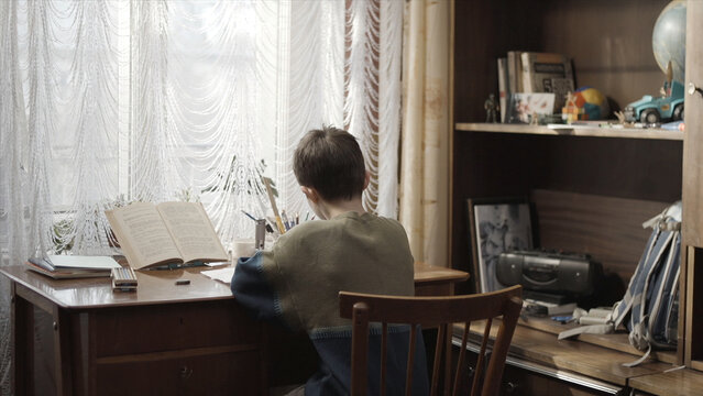 Rear view of boy doing homework sitting at desk at home. Reading book and doing homework. Young boy doing his homework - education concept