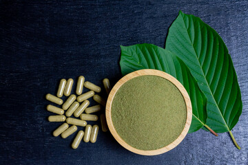 Mitragyna Speciosa Korth or kratom powder on wooden bowl wtih capsules and green leaf on black wooden background, top view and copy space. 