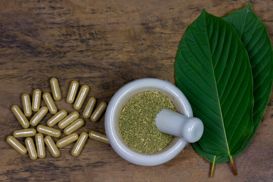 Mitragyna Speciosa Korth or kratom capsules with white mortar and pestle and green leaf on rustic wooden table, top view, copy space.