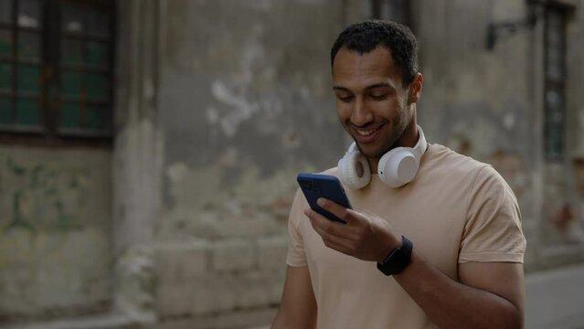 Side view of the african american runner walks down the street and records a voice message wearing the headphones and smart watch in his arm. Sport and healthy lifestyle concept.