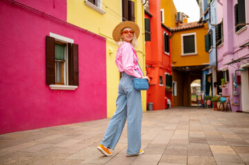 Happy smiling female traveler wearing stylish hat, glasses, pink shirt, wide leg trousers, walking,...