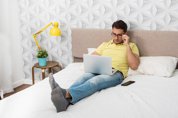 Thoughtful young businessman in casual wear lying on the bed and working on laptop. Distant job and...