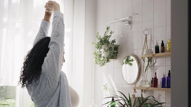 Young overweight woman stretching in light bathroom with minimalistic eco-flriendly design. White tiled wall with shadows from the window. Wellness and body positivity concept. 4K UHD