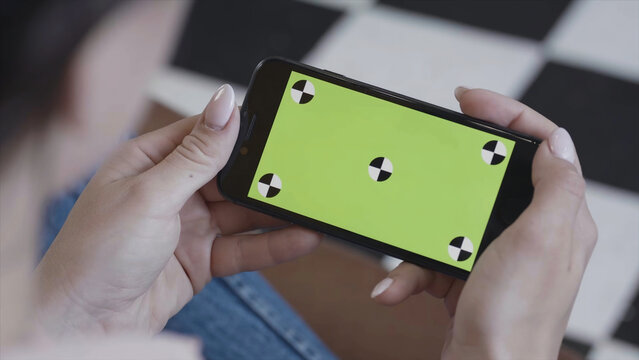 Female Teen Girl With Dark Hair Using Smartphone With Green Screen, Sitting Indoors. Stock Footage. Woman Holding Iphone With Chroma Key In Horizontal Position.