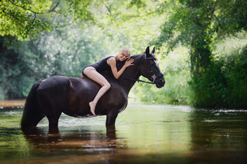 portrait of beautiful young blond woman laying on black horse in river in summer