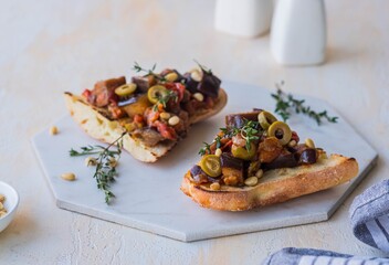 Sicilian caponata, eggplant, tomato and olive stew on white bread on a marble board against a light concrete background. Vegetable appetizer.