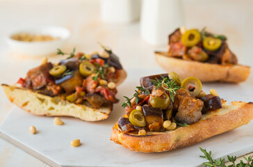Sicilian caponata, eggplant, tomato and olive stew on white bread on a marble board against a light concrete background. Vegetable appetizer.